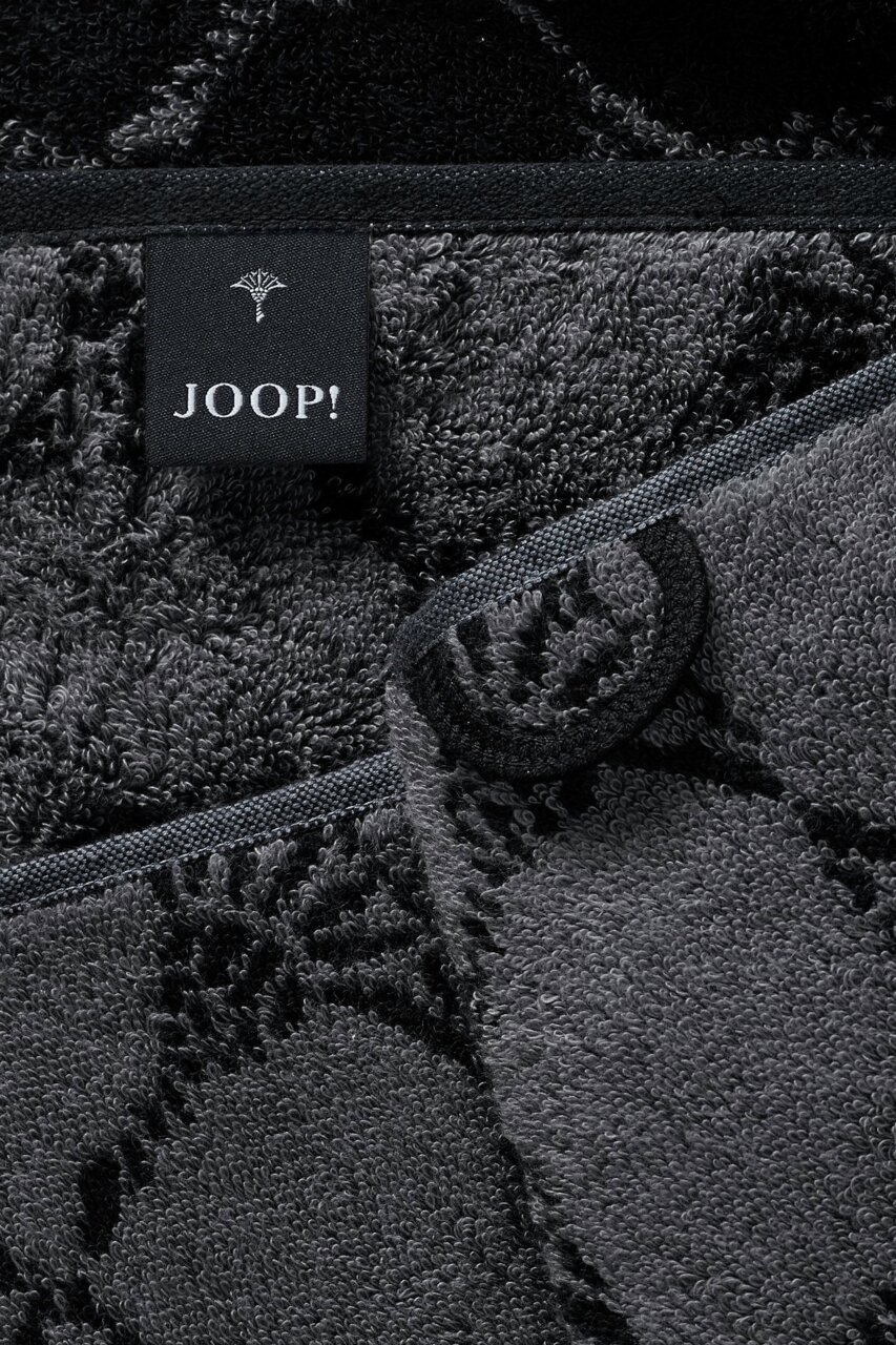 Close-up van een grijze saunadoek met een zwart label van het merk JOOP! en bloemmotieven.