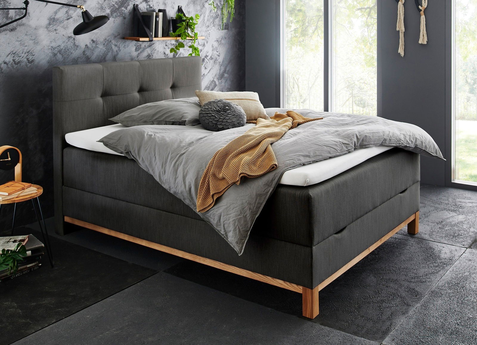 Modernes Boxspringbett in dunklem Stoff mit Holzfüßen, seitliche Perspektive, dekoriert mit grauer Bettwäsche und gelber Decke, in stilvollem Schlafzimmer mit großen Fenstern.