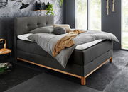 Modernes Boxspringbett in dunklem Stoff mit Holzfüßen, seitliche Perspektive, dekoriert mit grauer Bettwäsche und gelber Decke, in stilvollem Schlafzimmer mit großen Fenstern.