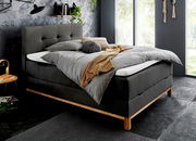 Modernes Boxspringbett in dunklem Stoff mit Holzfüßen, seitliche Perspektive, dekoriert mit grauer Bettwäsche und gelber Decke, in stilvollem Schlafzimmer mit großen Fenstern.