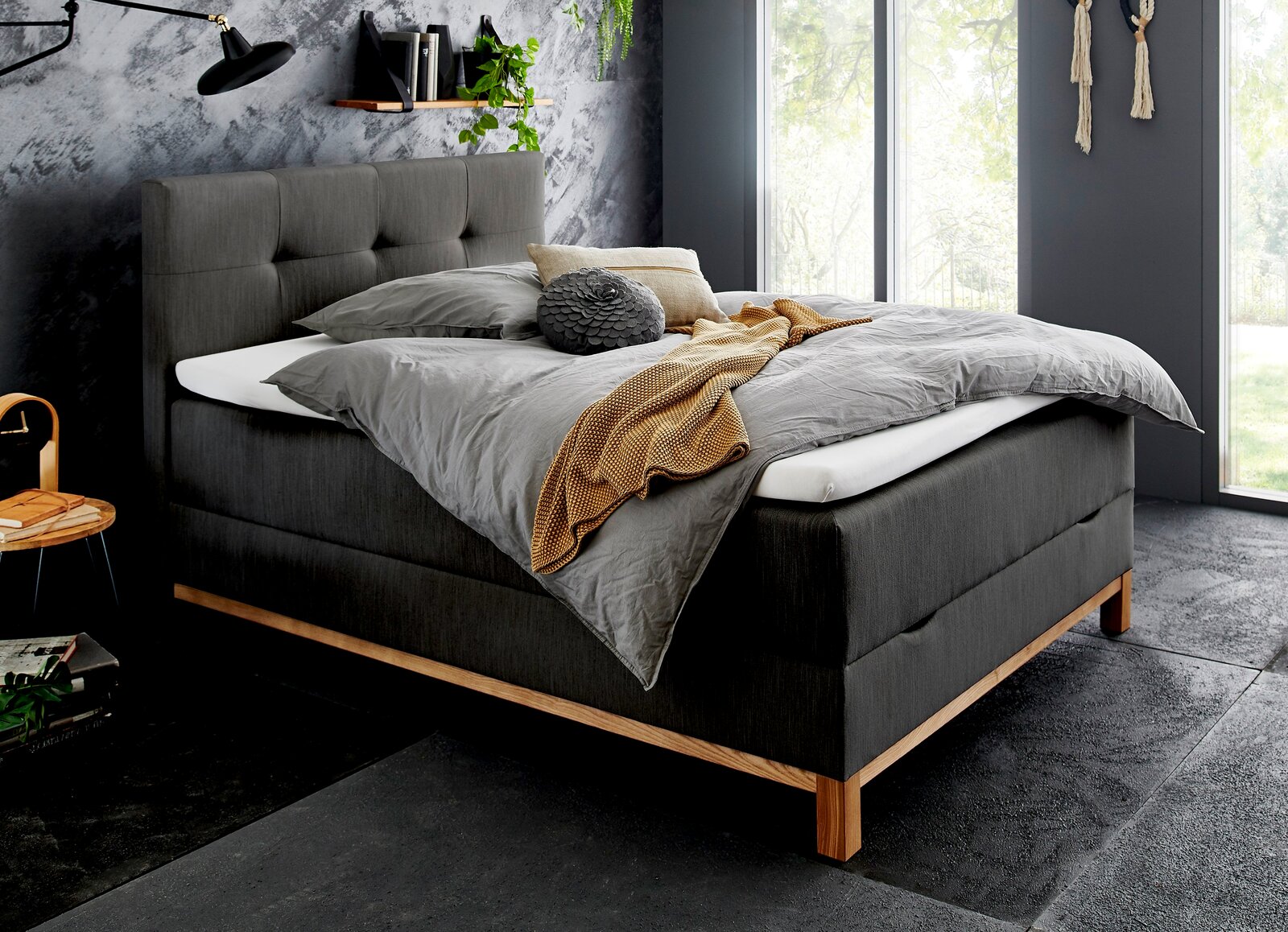 Modernes Boxspringbett in dunklem Stoff mit Holzfüßen, seitliche Perspektive, dekoriert mit grauer Bettwäsche und gelber Decke, in stilvollem Schlafzimmer mit großen Fenstern.