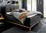 Modernes Boxspringbett in dunklem Stoff mit Holzfüßen, seitliche Perspektive, dekoriert mit grauer Bettwäsche und gelber Decke, in stilvollem Schlafzimmer mit großen Fenstern.