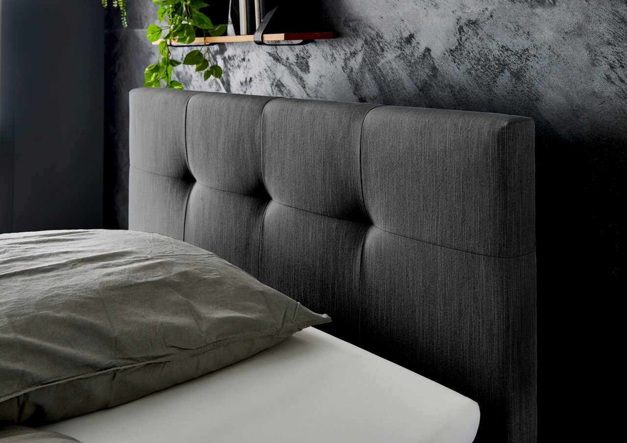 Nahaufnahme eines grauen Boxspringbett-Kopfteil mit gepolstertem Design, seitliche Perspektive