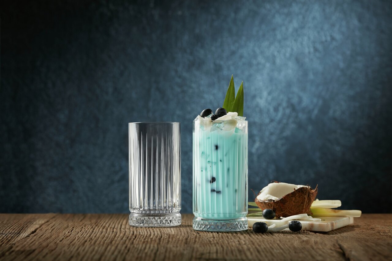 Zwei Longdrinkbecher Elysia auf einem Holztisch, einer leer und einer gefüllt mit einem blauen Getränk, garniert mit Beeren und Kokosnussstücken, seitliche Perspektive.