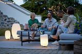Eine Gruppe von Menschen sitzt entspannt im Freien auf einem Sofa, umgeben von stilvollen Akku-Leuchten, die eine gemütliche Atmosphäre schaffen. Perspektive: seitlich von vorne.