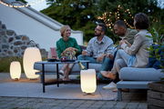 Eine Gruppe von Menschen sitzt entspannt im Freien auf einem Sofa, umgeben von stilvollen Akku-Leuchten, die eine gemütliche Atmosphäre schaffen. Perspektive: seitlich von vorne.