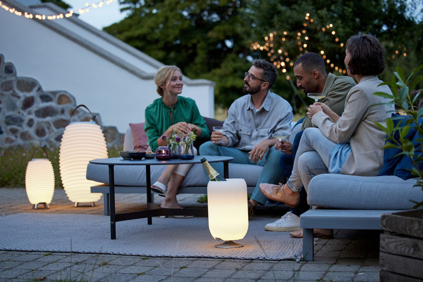 Eine Gruppe von Menschen sitzt entspannt im Freien auf einem Sofa, umgeben von stilvollen Akku-Leuchten, die eine gemütliche Atmosphäre schaffen. Perspektive: seitlich von vorne.