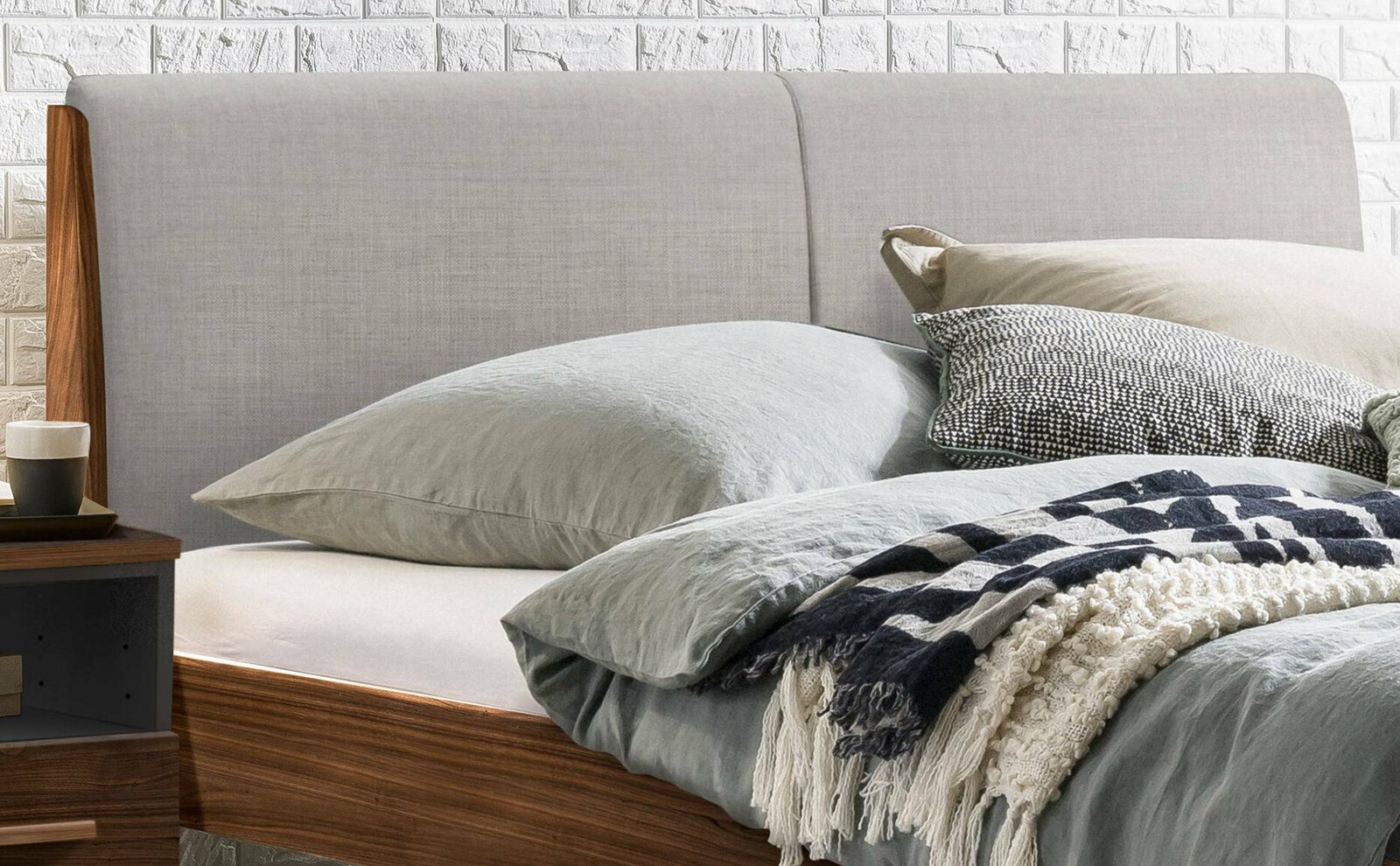 Seitliche Ansicht eines Bettgestells mit grauem Kopfteil, dekoriert mit Kissen und einer Decke.