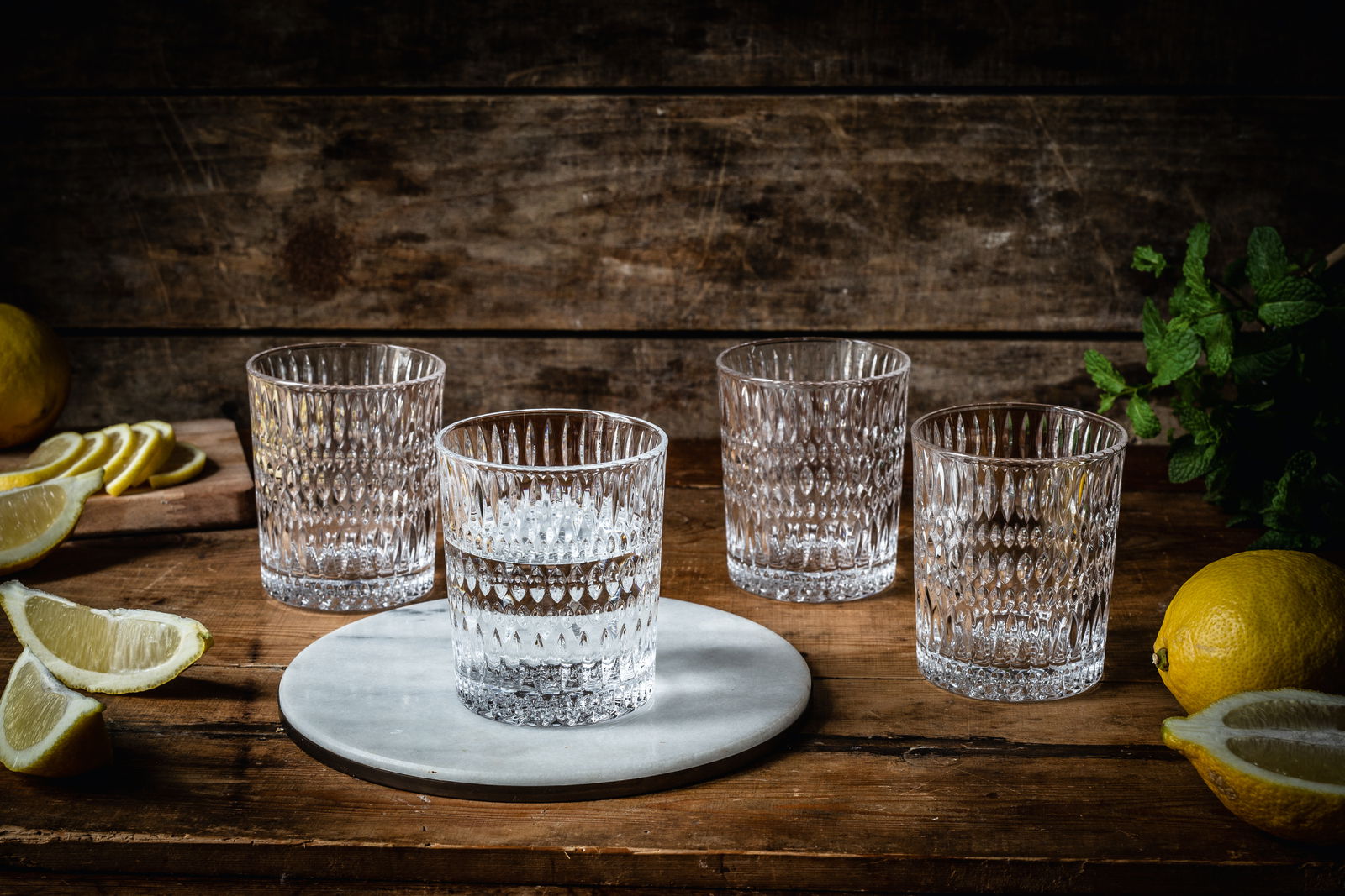Vier kristallklare Becher mit Ethno-Muster auf einem rustikalen Holztisch, umgeben von Zitronenscheiben und frischen Kräutern, aus einer leicht erhöhten Perspektive fotografiert.