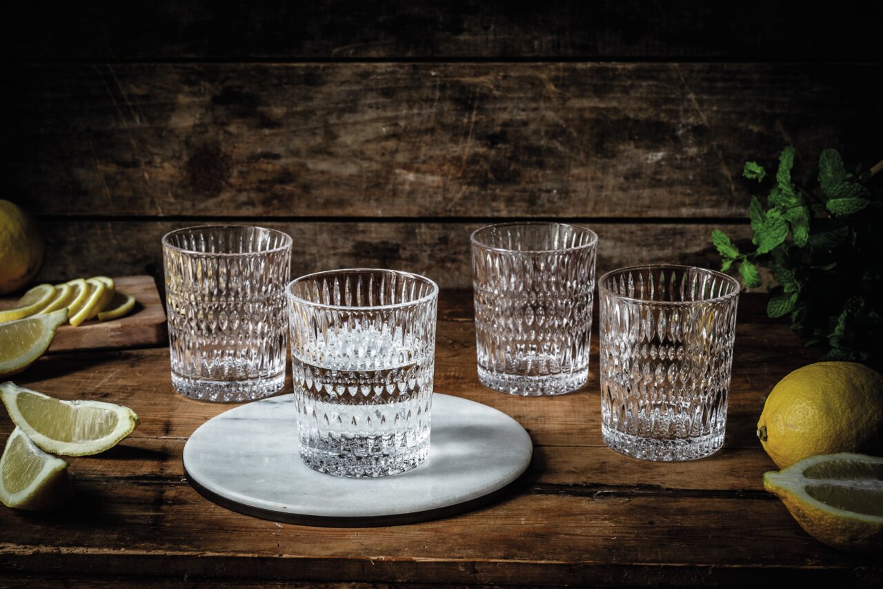 Vier kristallklare Becher mit Ethno-Muster auf einem rustikalen Holztisch, umgeben von Zitronenscheiben und frischen Kräutern, aus einer leicht erhöhten Perspektive fotografiert.