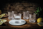 Vier kristallklare Becher mit Ethno-Muster auf einem rustikalen Holztisch, umgeben von Zitronenscheiben und frischen Kräutern, aus einer leicht erhöhten Perspektive fotografiert.