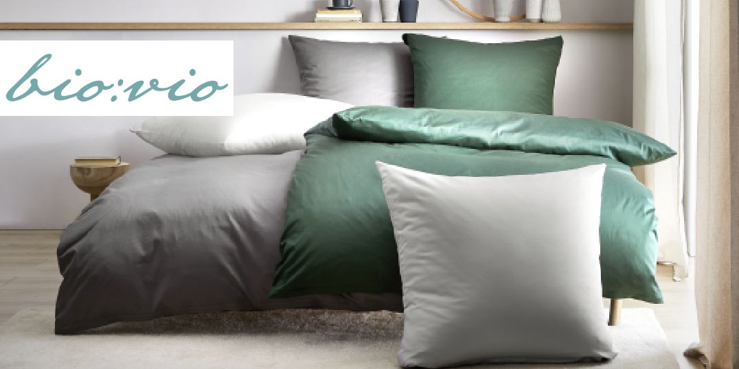Op de foto is een bed te zien met elegant grijs en donkergroen beddengoed en een bio vio-logo.