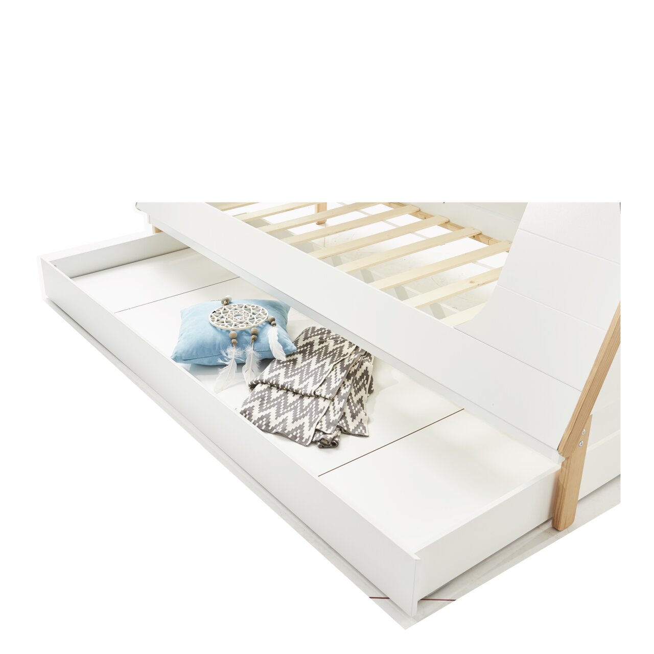 Een witte lade of een logeerbed onder een bedframe, met daarin een blauw kussen, een dromenvanger en een grijze deken. Aanzicht van bovenaf.