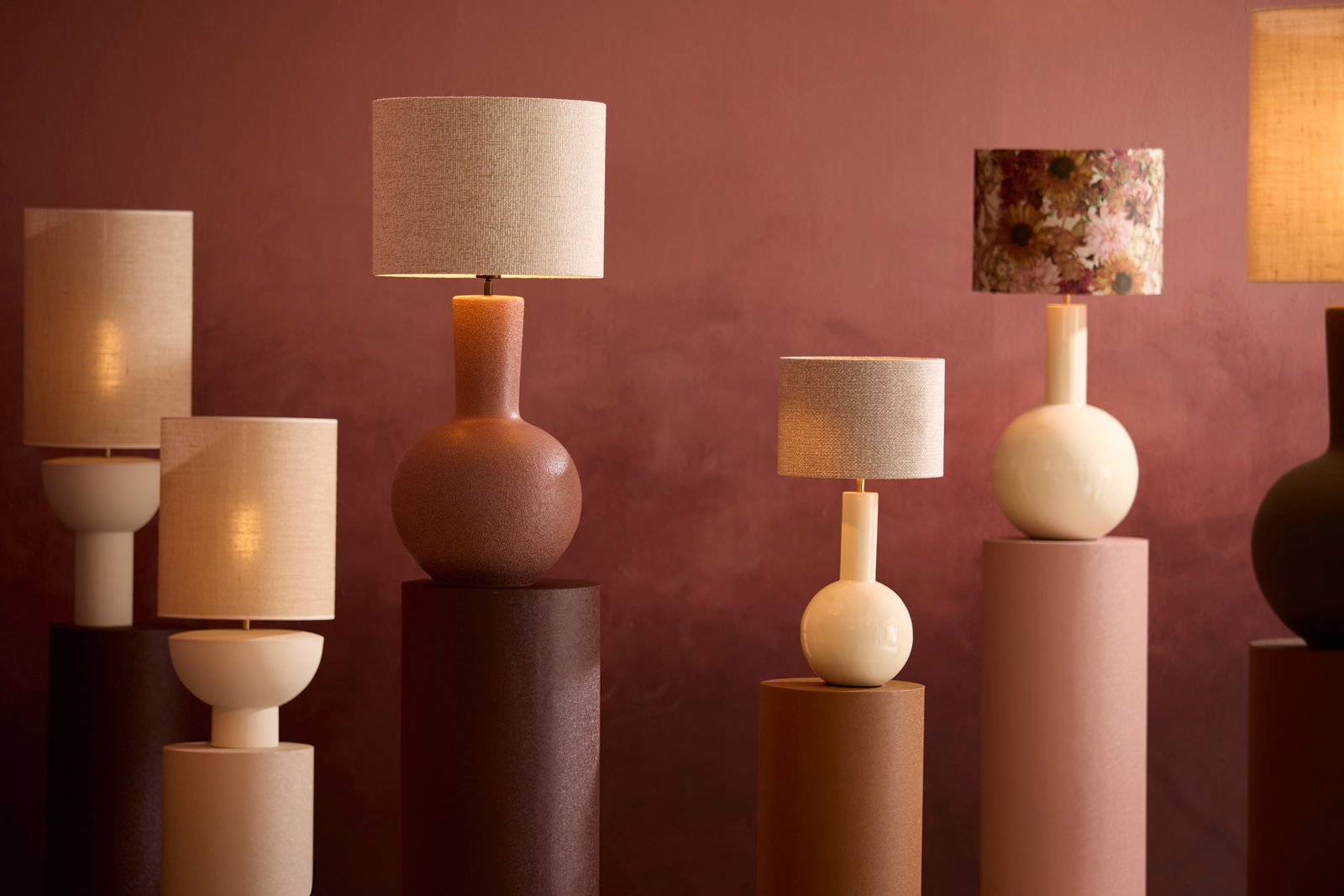 Verschiedene stilvolle Lampenschirme auf Sockeln vor einem warmen, roten Hintergrund, seitliche Perspektive.