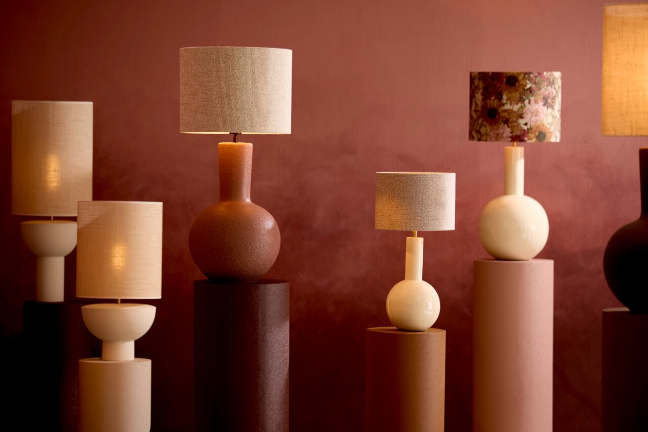 Verschiedene stilvolle Lampenschirme auf Sockeln vor einem warmen, roten Hintergrund, seitliche Perspektive.