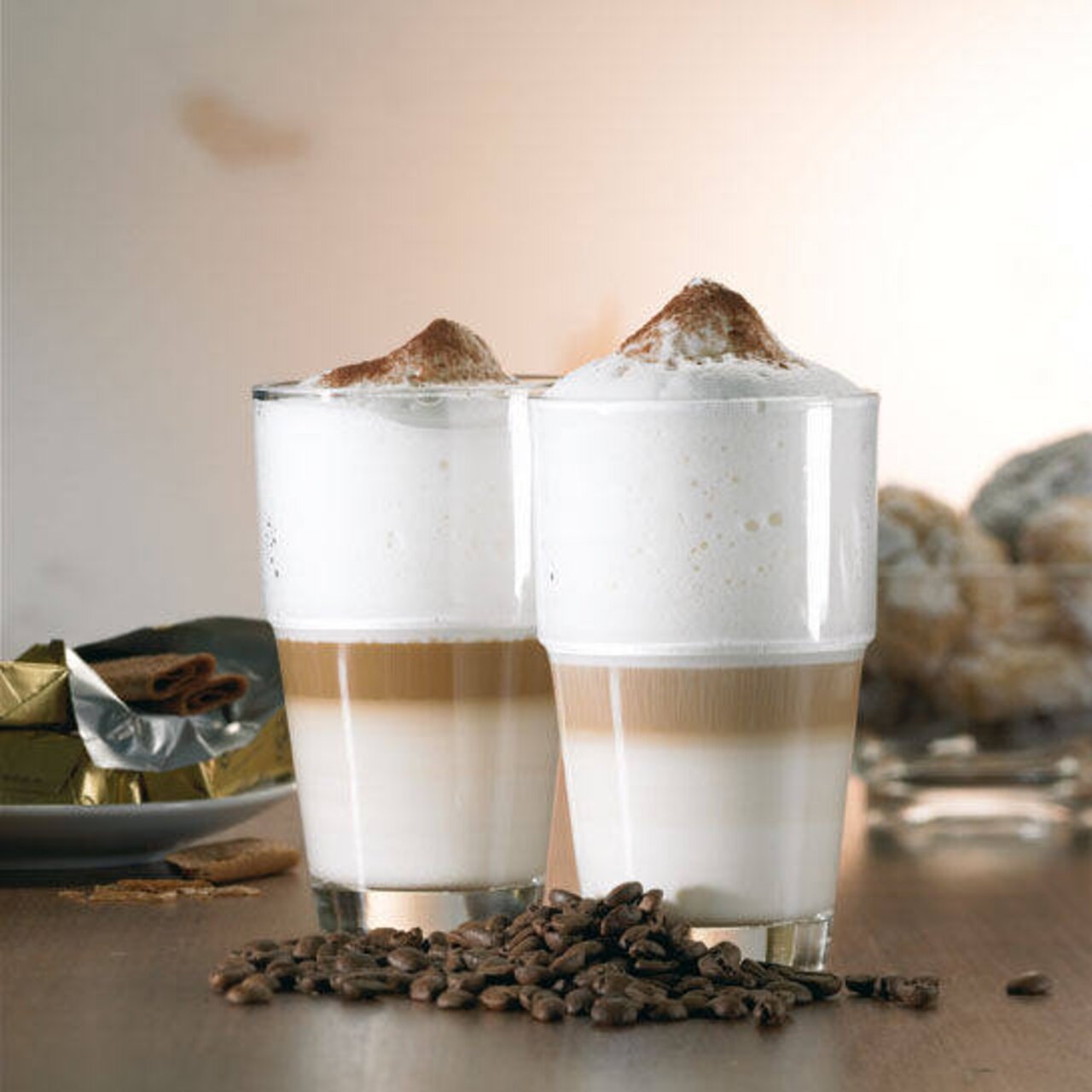 Zwei Latte Macchiato in hohen Gläsern mit Milchschaum und Kakaopulver, umgeben von Kaffeebohnen, aus seitlicher Perspektive fotografiert.