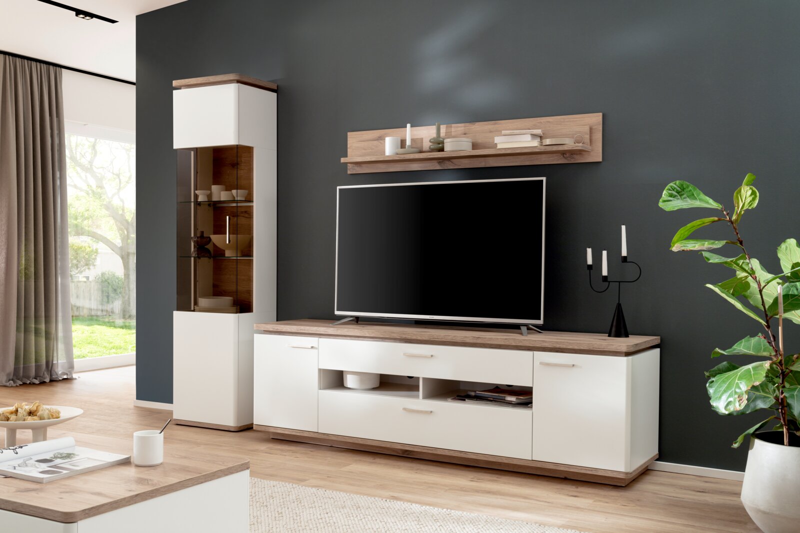 Modernes TV-Wohnwand-Set in Weiß und Holzoptik mit Lowboard, Vitrine und Wandboard vor dunkelgrauer Wand, Perspektive: frontale Ansicht leicht von links