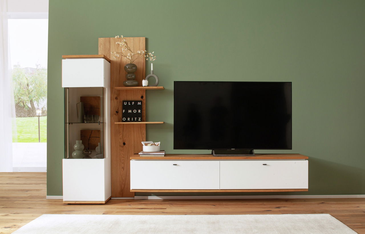 Frontale Ansicht der Wandkombination Novel MATADI mit weißem und holzfarbenem Design, bestehend aus einem TV-Schrank, einem Vitrinenschrank und Regalböden, vor einer grünen Wand.