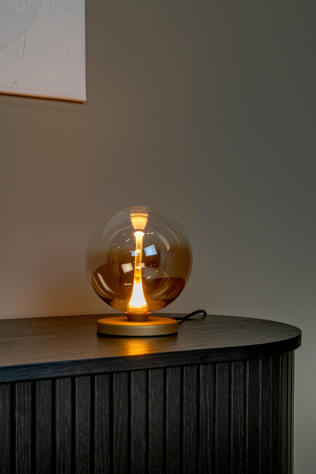 Leuchtende Tischlampe mit transparenter Glaskugel und warmweißem LED-Filament auf rundem Sockel, frontal aus leicht erhöhter Perspektive auf einem dunklen Sideboard vor grauer Wand