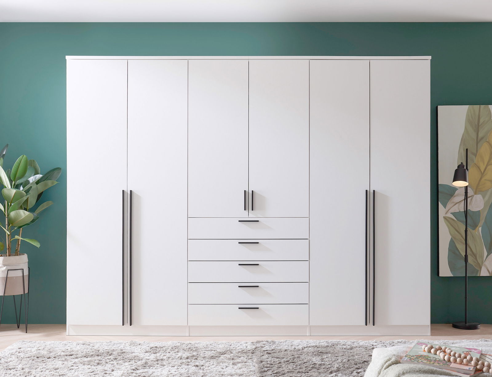 6-türiger Drehtürenschrank in Weiß mit schwarzen Griffen, frontal fotografiert, vor einer grünen Wand mit Pflanzen und Dekoration.