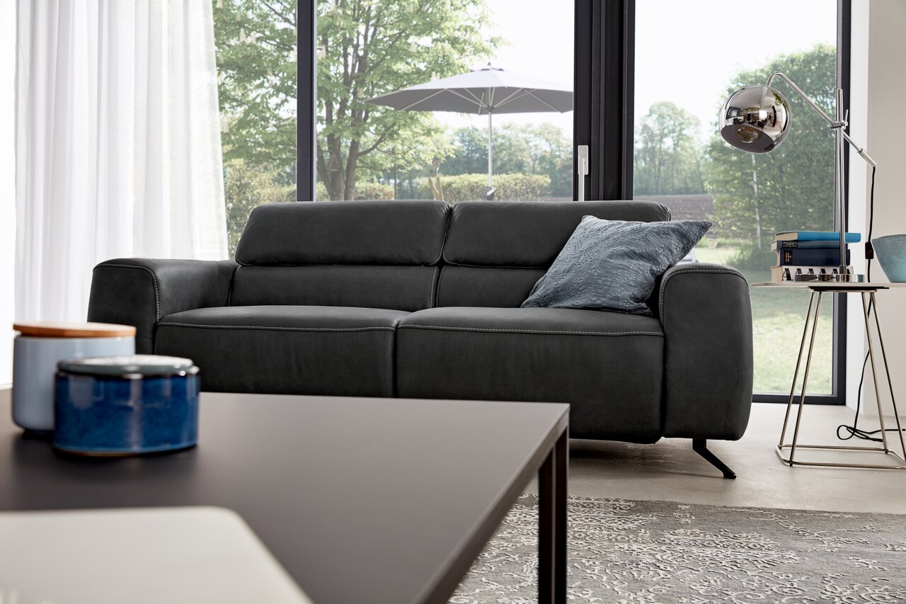 Musterring Sofa 3er MR 260 Modernes, dunkelgraues 3-Sitzer-Sofa in einem hellen Wohnzimmer mit Blick auf den Garten durch große Fenster.
