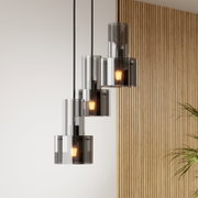 Moderne Pendelleuchte mit drei rauchgrauen Glaszylindern und warm leuchtenden Filament-Leuchtmitteln, an schwarzen Kabeln hängend, in leichter Seitenansicht vor heller Wand mit Holzlamellen