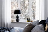 Schwarze Tischleuchte LOFTY auf einer Fensterbank, umgeben von Büchern und Dekoration, mit Blick auf eine verschneite Waldlandschaft.