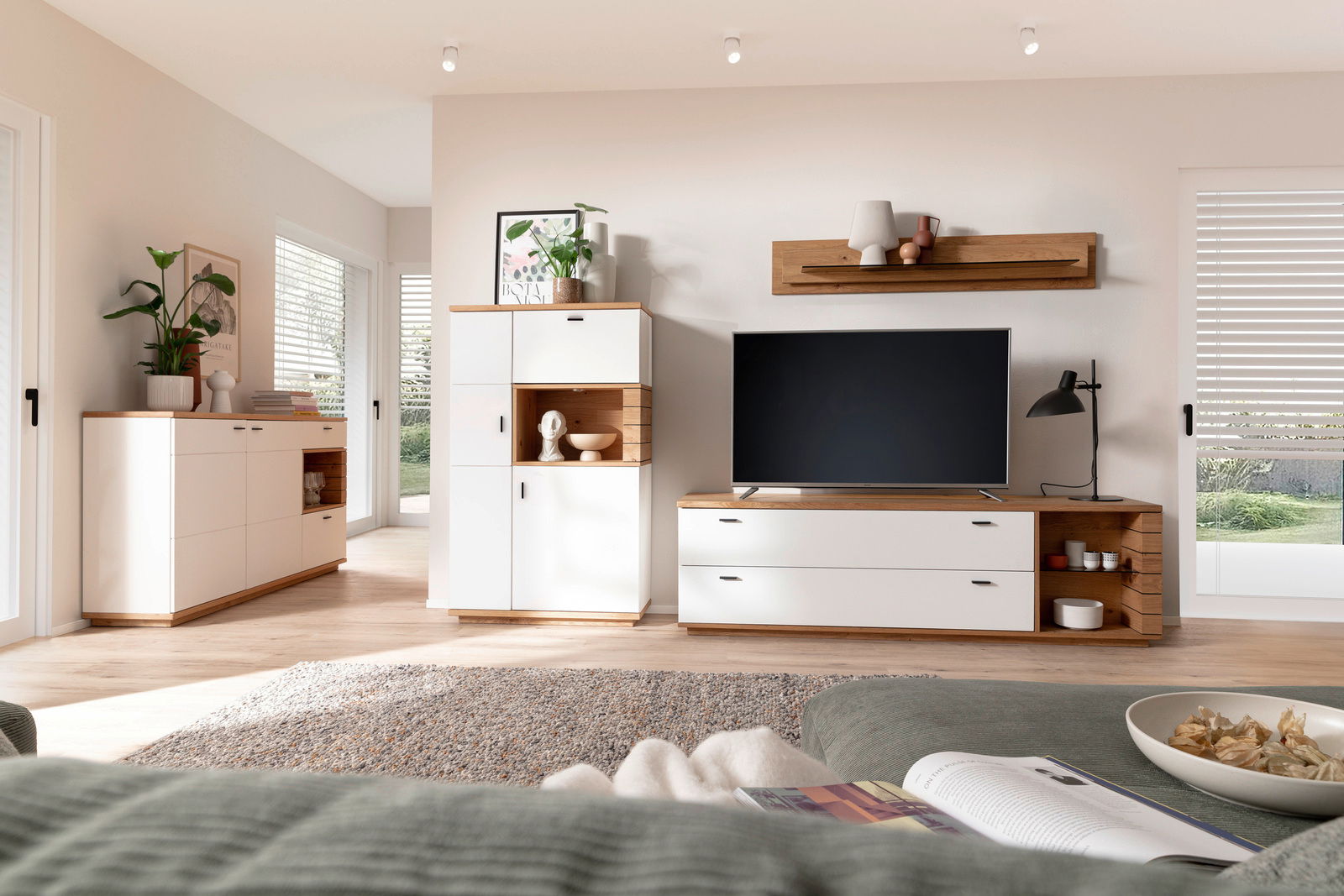 Novel Highboard MANITOBA Modernes Highboard in einem hellen Wohnzimmer, frontal fotografiert, mit weißen und holzfarbenen Elementen, dekoriert mit Pflanzen und Kunstobjekten.