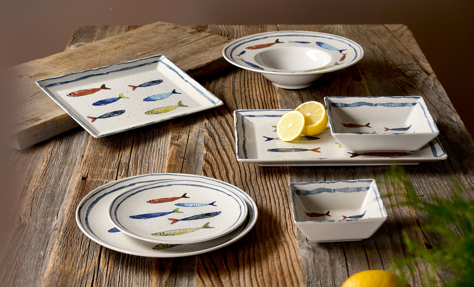 Maritimes Geschirrset aus weißer Keramik mit farbenfrohem Fischdekor und blauen Randlinien, bestehend aus Tellern, Platten und Schalen, dekorativ arrangiert auf einem rustikalen Holztisch in schräger Draufsicht.