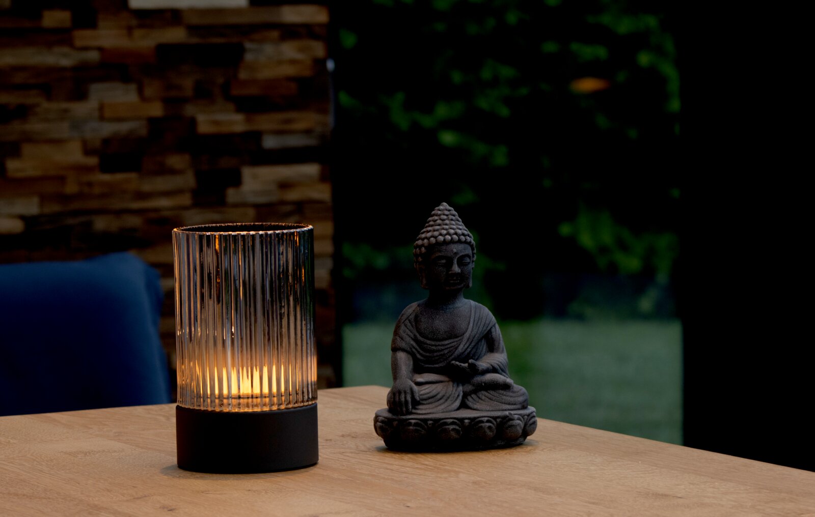 Stimmungsvolle Akku-Tischleuchte mit geriffeltem Glas, leuchtend auf einem Holztisch neben einer Buddha-Statue, seitliche Perspektive.