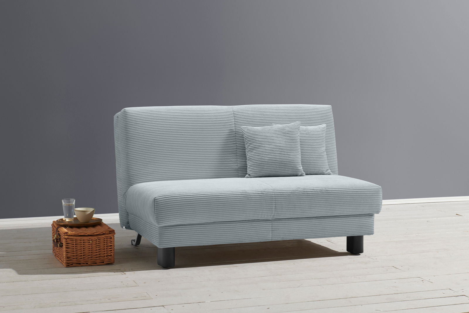 Graues Funktionssofa mit zwei Kissen, frontal fotografiert, auf hellem Holzboden neben einem kleinen Korbtisch mit Glas und Schale.
