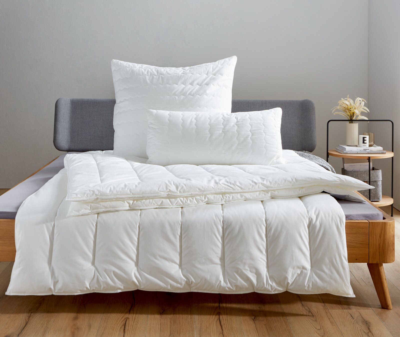 Het Centa Star Inspiration-bed voor alle seizoenen in een moderne slaapkamer, van voren gefotografeerd, met witte kussens en een deken op een grijs bed.