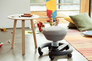 Kinderzimmer mit einem grauen Sitzhocker auf Rollen und einem Holztisch, seitliche Perspektive.