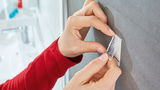 Nahaufnahme einer Person, die einen tesa Powerstrip Waterproof an einer grauen Wand im Badezimmer anbringt.