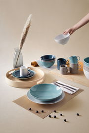 Verschiedene Geschirrteile in Blau- und Grautönen, darunter Tassen, Schalen und Teller, auf einem Tisch arrangiert. Eine Hand hält eine weiße Schale. Perspektive von oben.