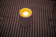 Geriffelte weiße Kerze im Glas mit warm gelb leuchtender Flamme auf gestreiftem Untergrund, leicht von oben fotografiert.