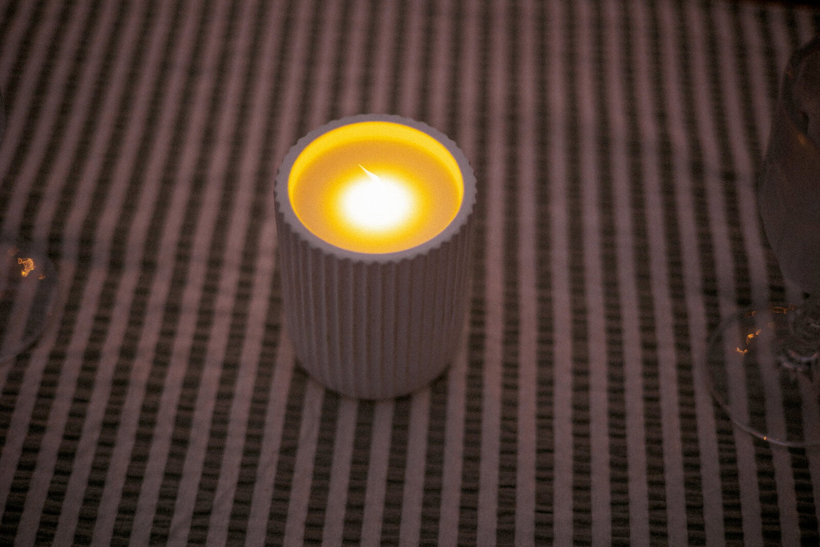 Een geribbelde witte kaars in een glas met een warmgele vlam op een gestreepte achtergrond, licht van bovenaf gefotografeerd.