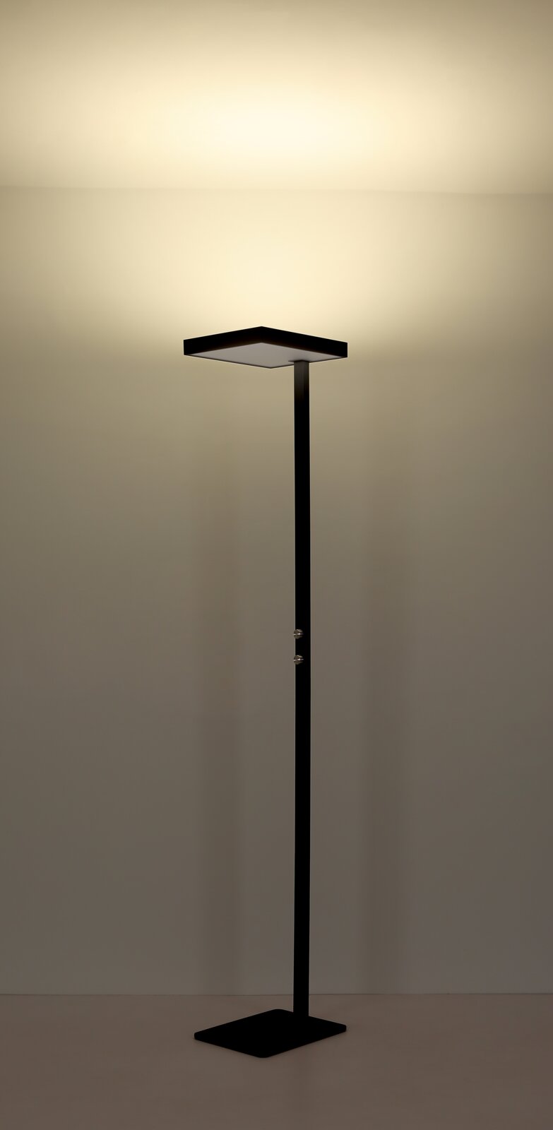 Schwarze, minimalistische LED-Stehleuchte mit quadratischem Leuchtenkopf und schlankem Stab auf quadratischem Standfuß, frontal in leichter Untersicht fotografiert, warmes indirektes Licht nach oben an die Wand.