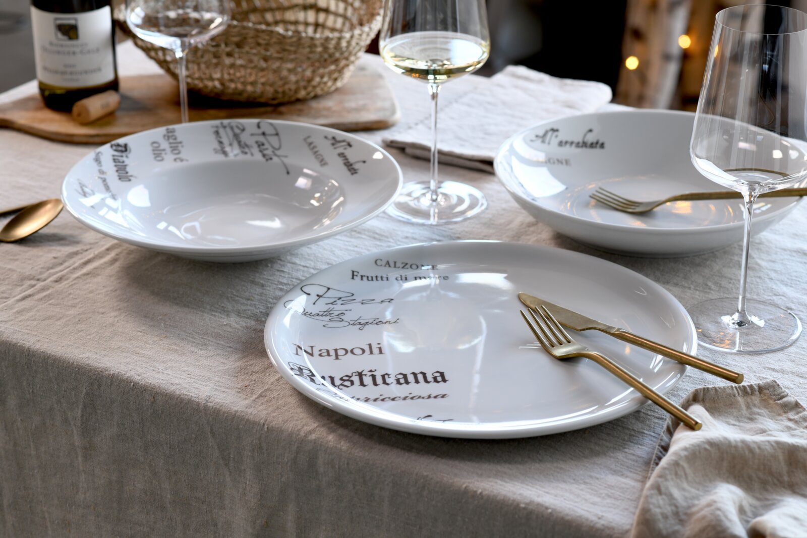 Gedeckter Tisch mit weißem Porzellan-Geschirr mit italienischem Schriftzug (Teller und tiefe Schalen), goldfarbenem Besteck und Weingläsern; Perspektive: leicht erhöhte, schräge Ansicht von vorn.