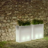 Zwei leuchtende, rechteckige Blumentöpfe Junco 80 vor einer Steinmauer, mit grünen Pflanzen gefüllt, in einer nächtlichen Gartenszene. Perspektive von vorne.