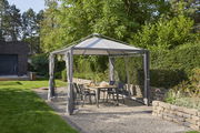 Gartenpavillon mit grauem Dach und Vorhängen, umgeben von grüner Hecke und Bäumen, mit Tisch und Stühlen darunter, seitliche Perspektive