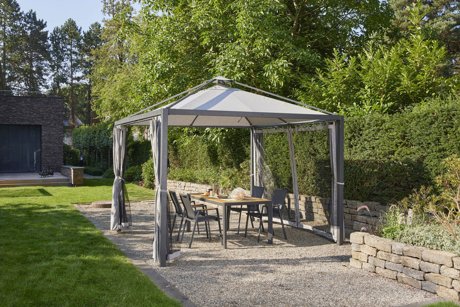 Gartenpavillon mit grauem Dach und Vorhängen, umgeben von grüner Hecke und Bäumen, mit Tisch und Stühlen darunter, seitliche Perspektive