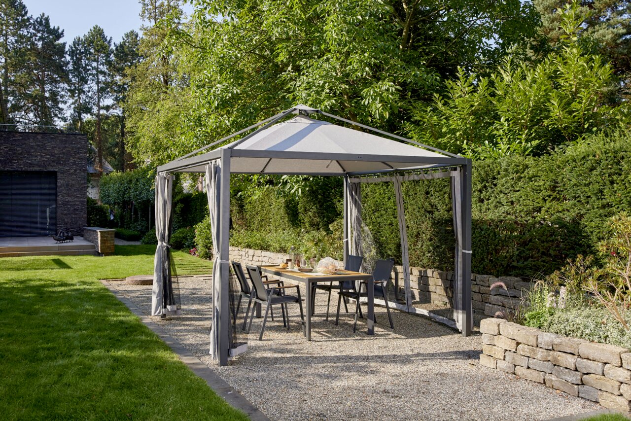 Gartenpavillon mit grauem Dach und Vorhängen, umgeben von grüner Hecke und Bäumen, mit Tisch und Stühlen darunter, seitliche Perspektive