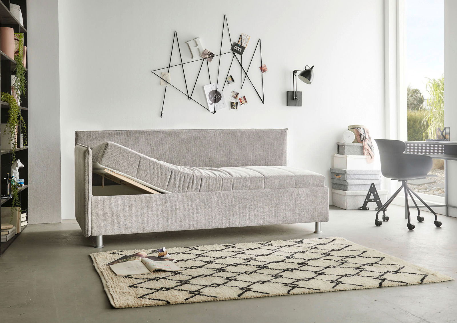 Graue Polsterliege in modernem Wohnzimmer, seitliche Perspektive, mit hochgeklappter Liegefläche und Stauraum darunter.