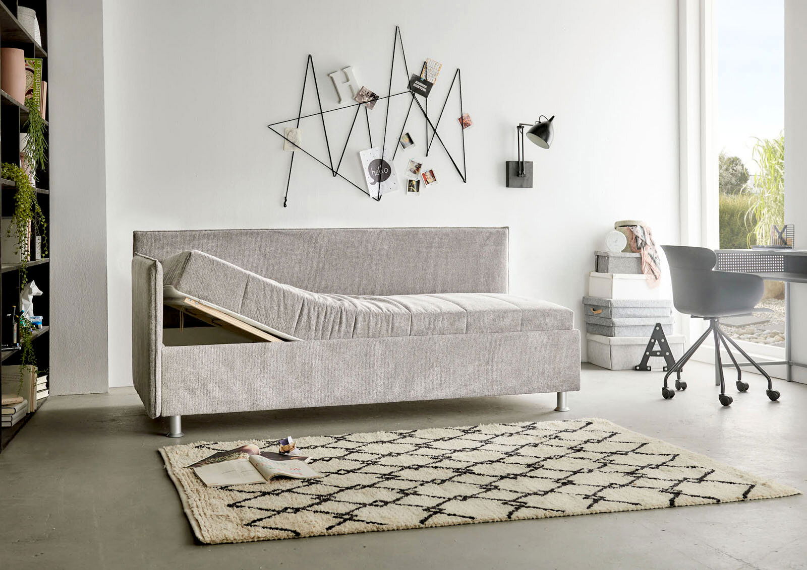 Graue Polsterliege in modernem Wohnzimmer, seitliche Perspektive, mit hochgeklappter Liegefläche und Stauraum darunter.