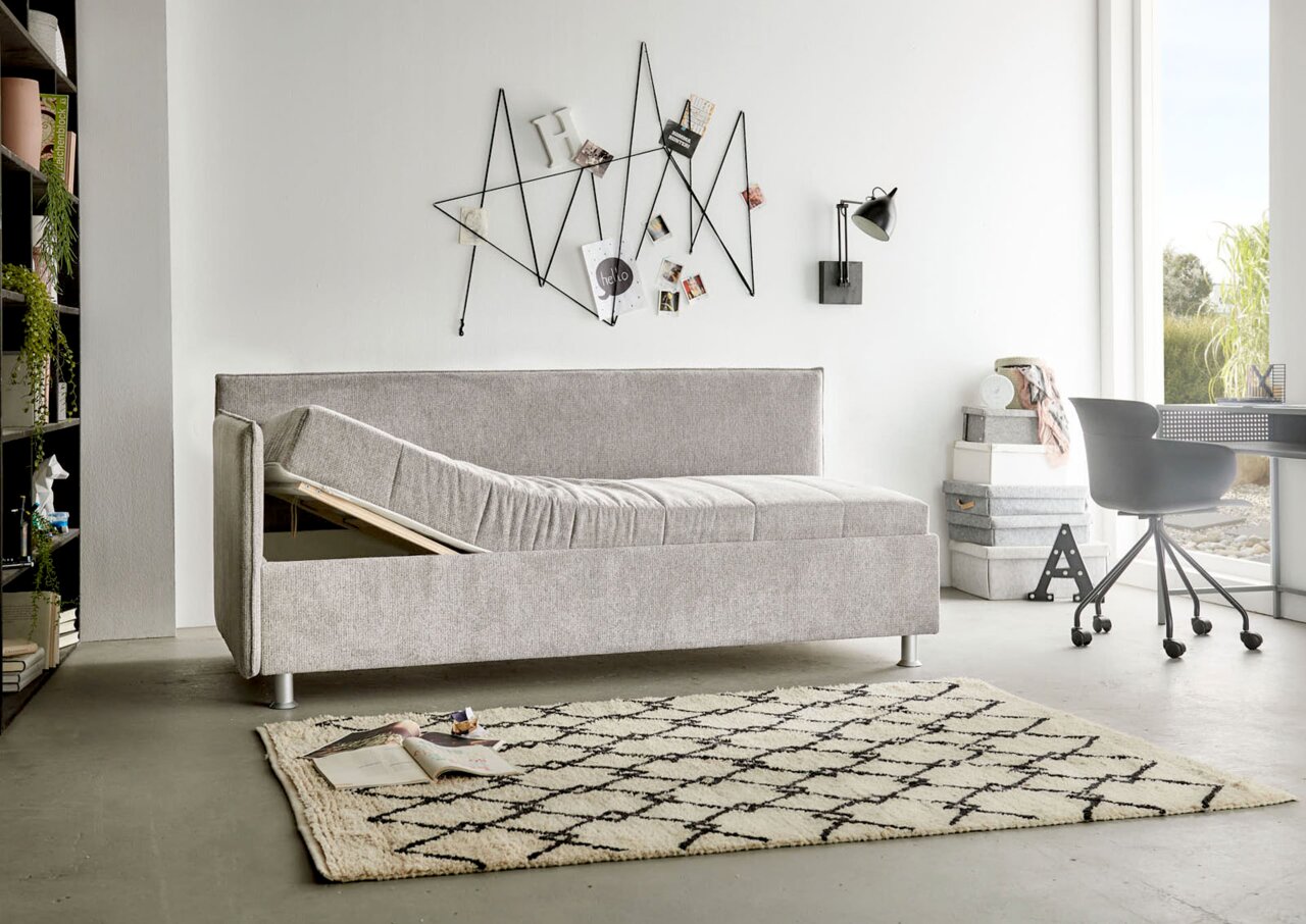 Graue Polsterliege in modernem Wohnzimmer, seitliche Perspektive, mit hochgeklappter Liegefläche und Stauraum darunter.