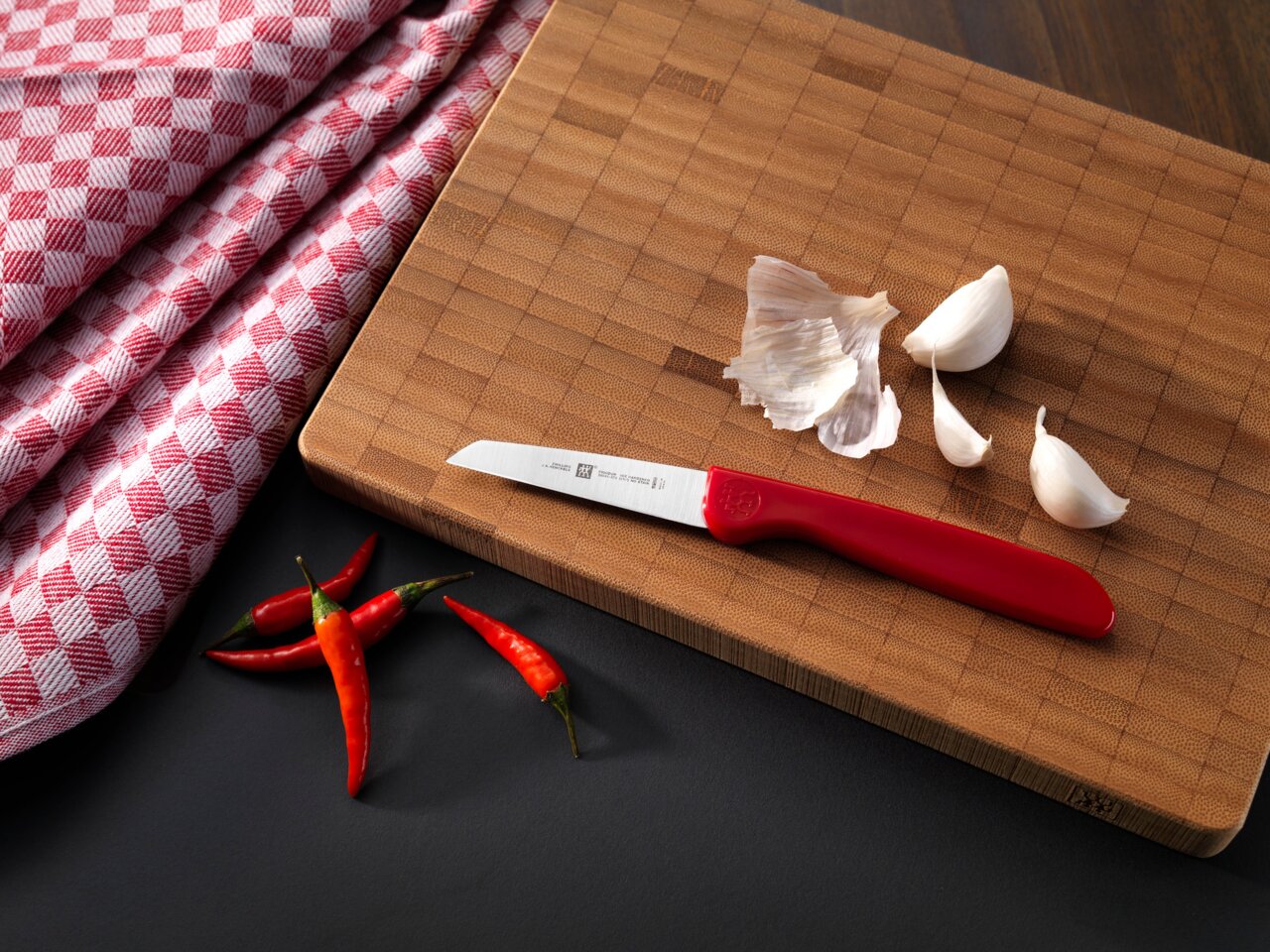 ZWILLING Gemüsemesser 7cm Gemüsemesser mit rotem Griff auf einem Holzbrett, umgeben von Knoblauchzehen und roten Chilischoten, aus der Vogelperspektive.