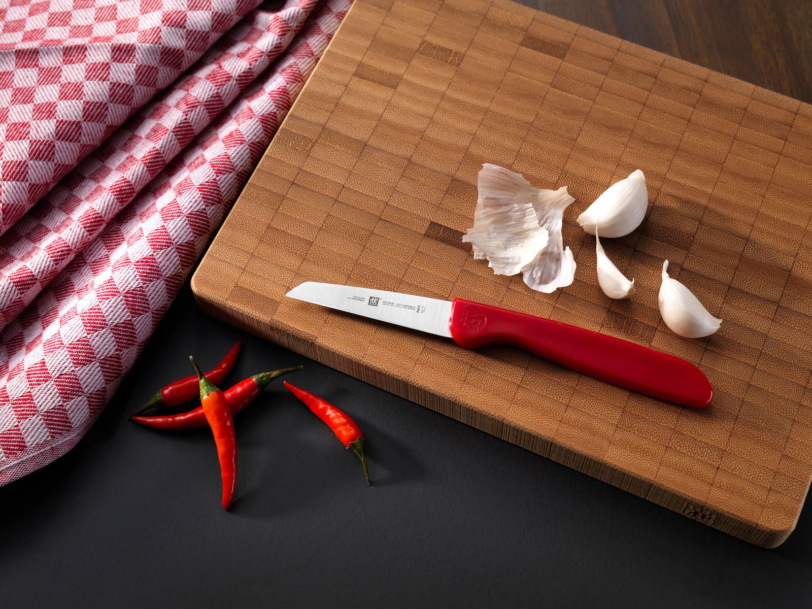 Gemüsemesser mit rotem Griff auf einem Holzbrett, umgeben von Knoblauchzehen und roten Chilischoten, aus der Vogelperspektive.