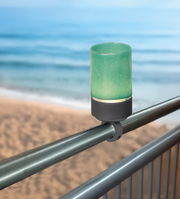 Geländerhalterung mit grünem Akku-Windlicht, montiert an einem Metallgeländer mit Blick auf das Meer im Hintergrund.