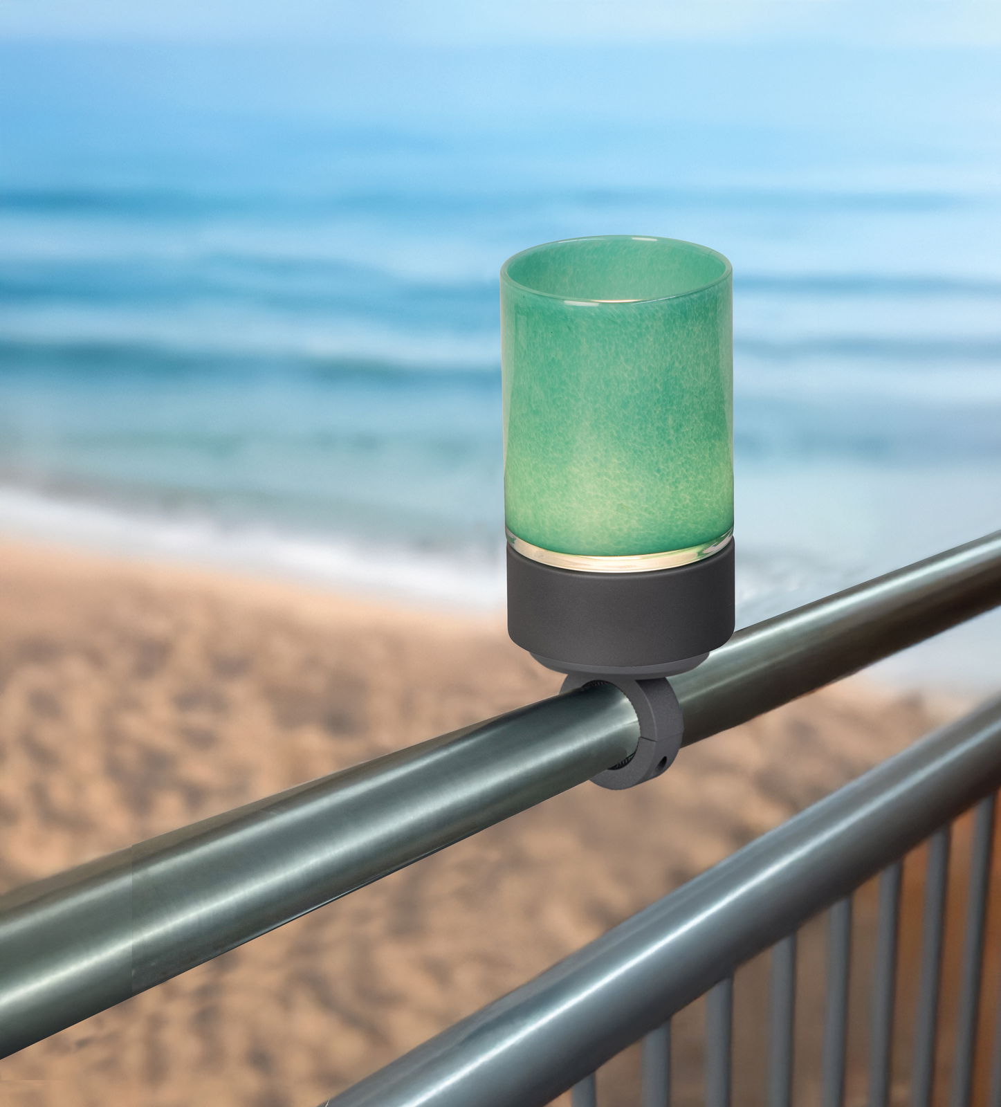 Geländerhalterung mit grünem Akku-Windlicht, montiert an einem Metallgeländer mit Blick auf das Meer im Hintergrund.